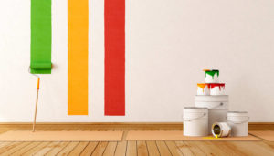 Botes de pintura apilados sobre suela de parket, pared blanca con tres franjas de colores pintadas con rodillo